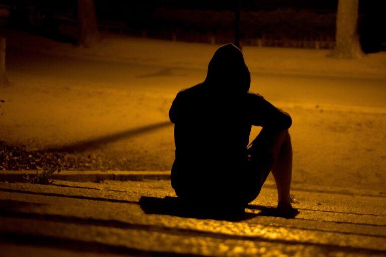 Man with hoodie sitting alone on the street in the dark underneath a streetlight